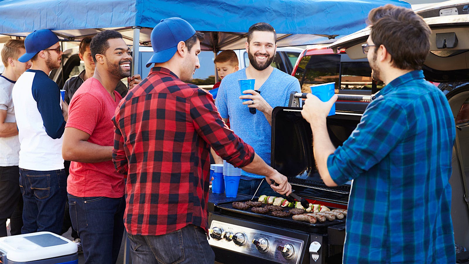 Men tailgating, standing around a grill drinking out of plastic cups