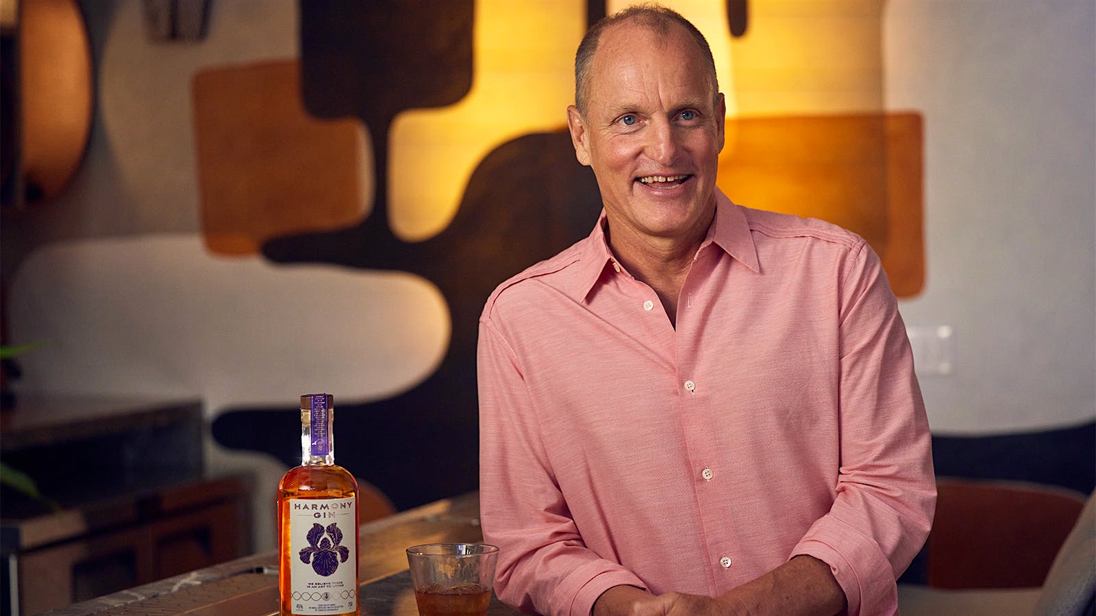 Woody Harrelson leaning on a bar next to a bottle and glass of Harmony gin