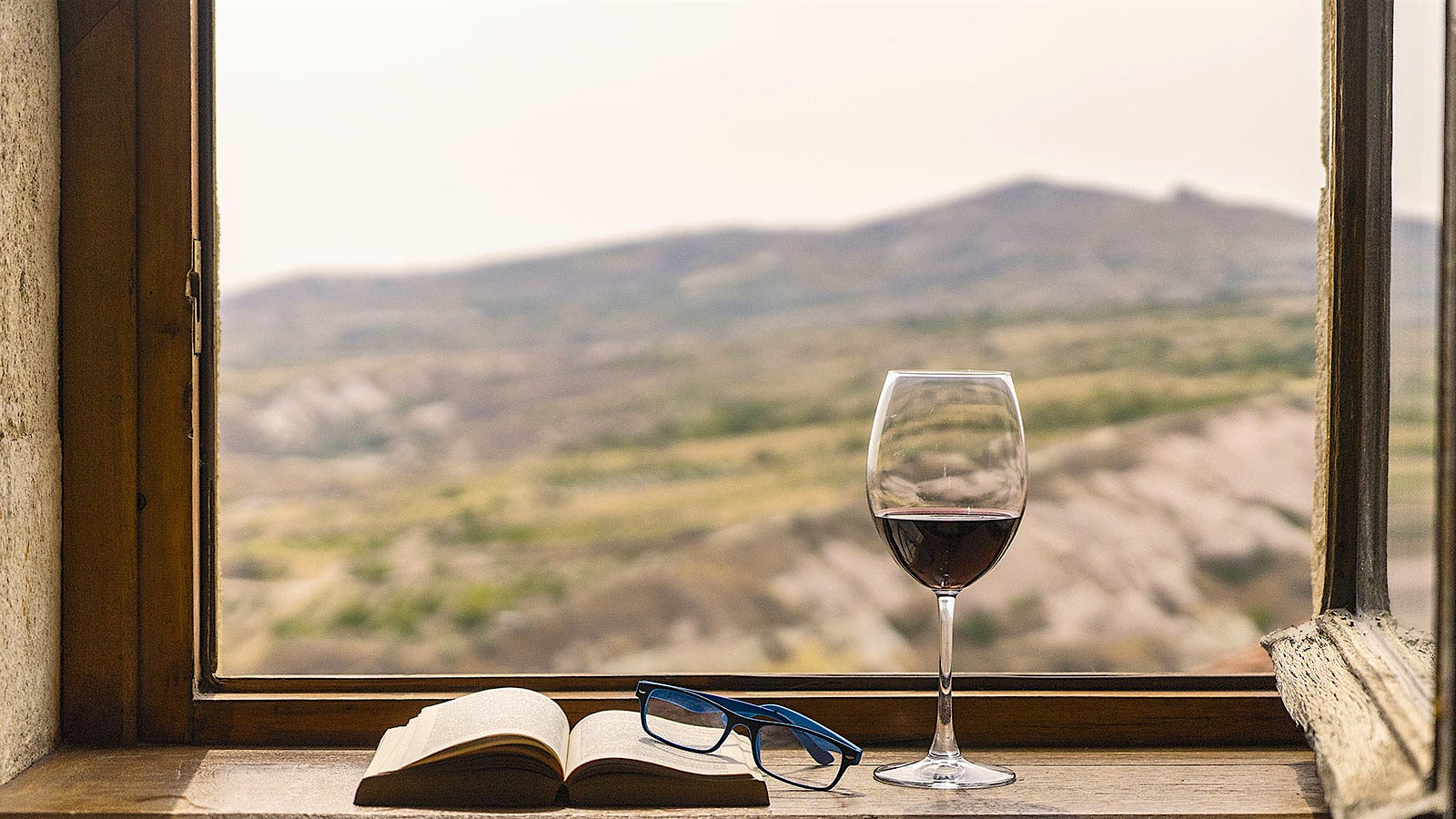 A glass of red wine, an open book and a pair of glasses on a ledge in front of a window looking out onto an empty sloping field with a mountain in the background.